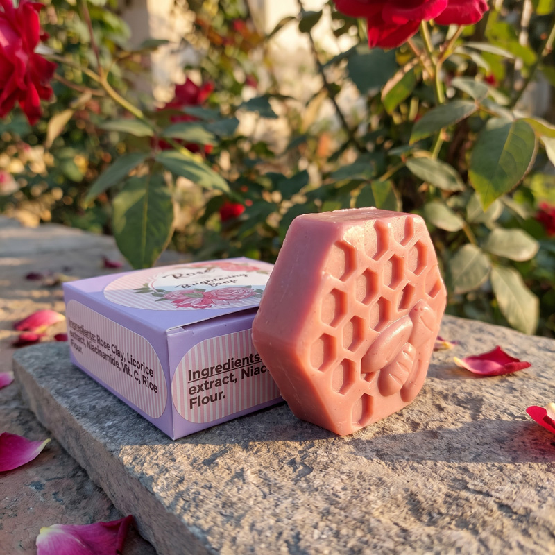 Zenvio Naturals Rose Brightening Soap with Ingredients Box Close-up of Zenvio Naturals hexagonal Rose Brightening Soap with a bee and honeycomb design leaning against its light purple packaging box on a stone surface.