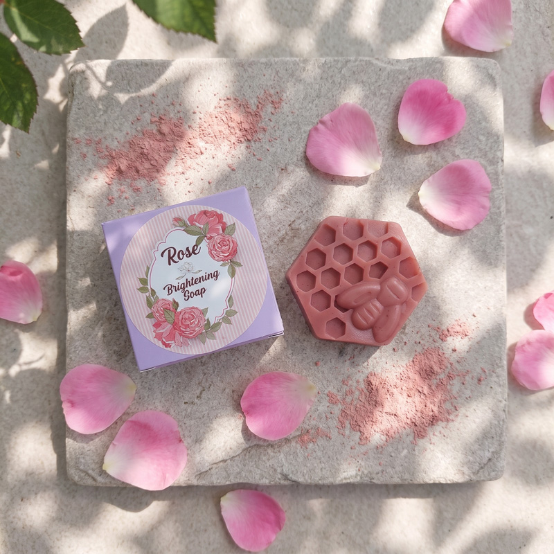 Top-Down View of Rose Brightening Soap and Box with Petals Flat lay top-down view of Zenvio Naturals Rose Brightening Soap and its floral-labeled box on a light stone tile, surrounded by scattered pink rose petals and rose clay powder under dappled sunlight.